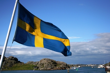 Pictured is the Swedish flag flying from the stern of a ship; the Swedish landscape provides a lovely backdrop.